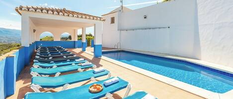A pool area with blue lounge chairs and a white building with a tiled roof.