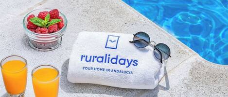 A towel with "ruralidays" and "your home in Andalucia" text, sunglasses, a bowl of raspberries, and two glasses of orange juice by a pool.