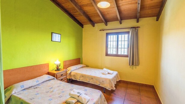A room with two beds, a nightstand with a lamp, a window with curtains, and a small picture on the wall. - Alejandro de Almachar Almáchar by Ruralidays (Málaga)