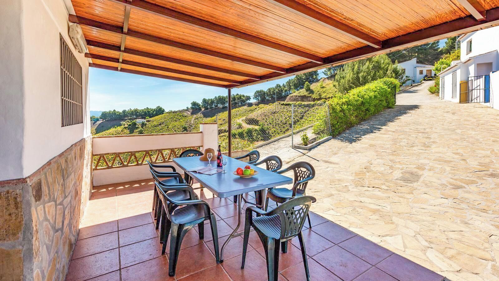 A covered outdoor patio with a table and chairs, a view of a vineyard, and a stone pathway leading to a building.