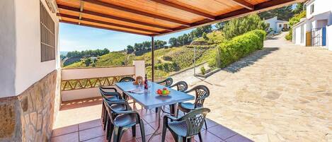 A covered outdoor patio with a table and chairs, a view of a vineyard, and a stone pathway leading to a building.