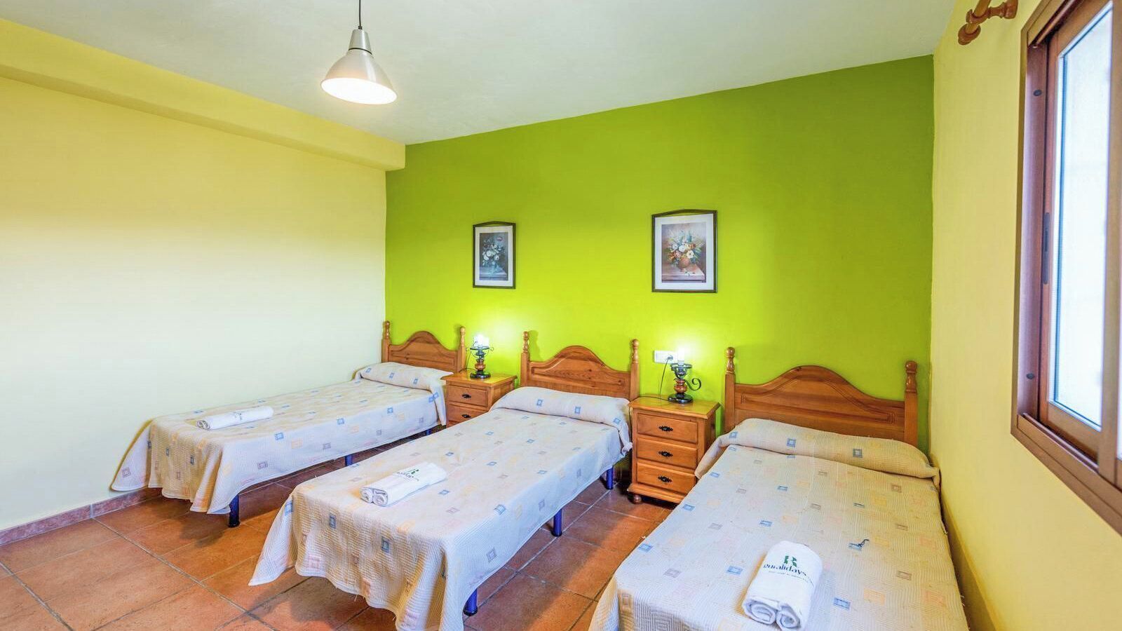 Een kamer met drie eenpersoonsbedden, elk met een houten hoofdbord en een nachtkastje met een lamp. De muren zijn geverfd in twee kleuren, geel en groen.