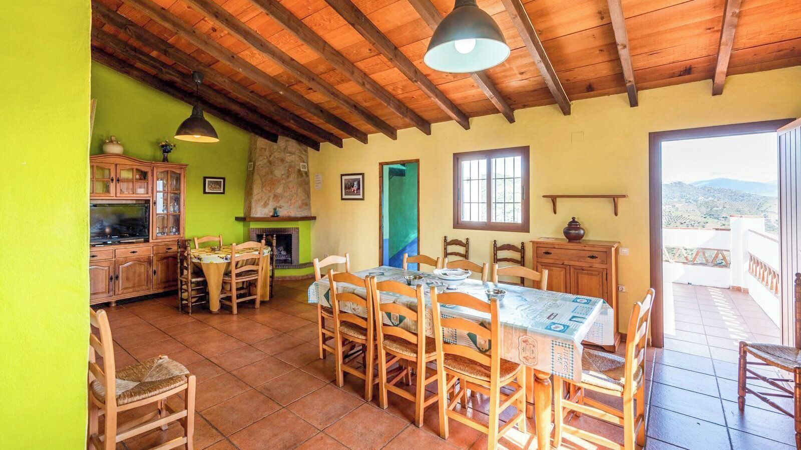 A dining area with a large table and chairs, a kitchen with cabinets and a stove, and a view of the outdoors through a door.