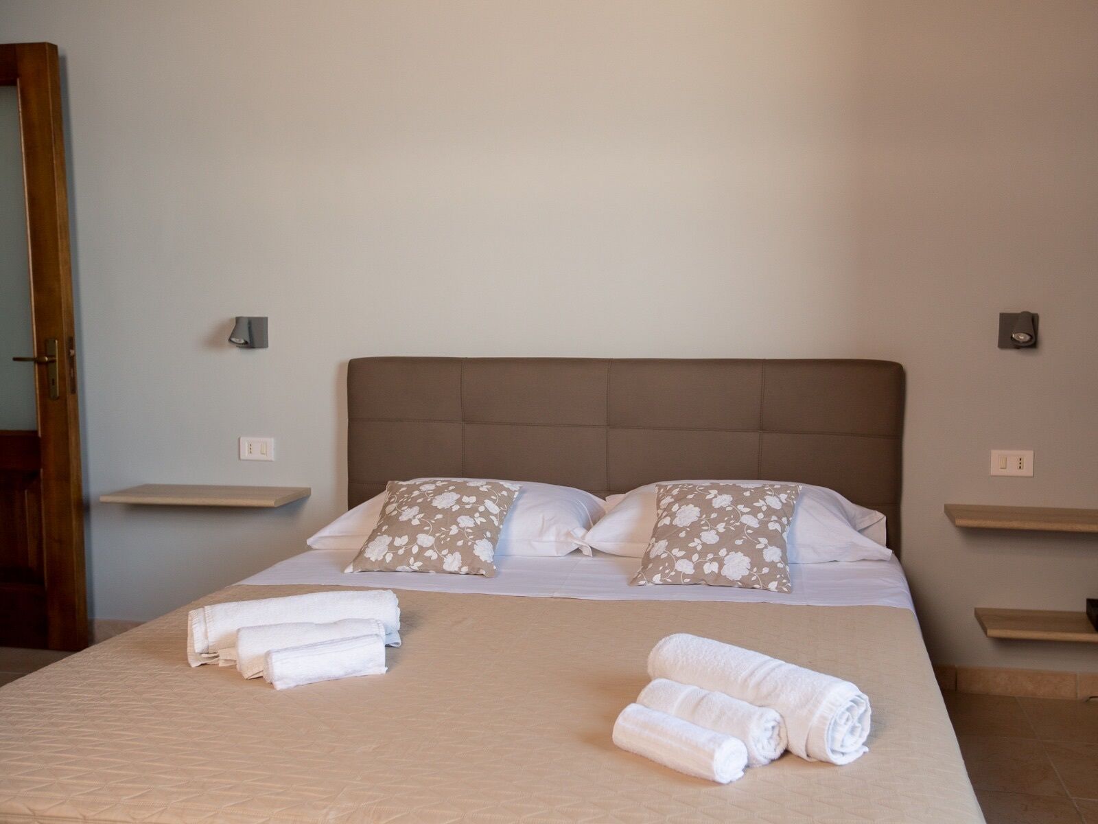 A neatly made bed with a brown headboard, two pillows with a floral pattern, and rolled towels on the bed.