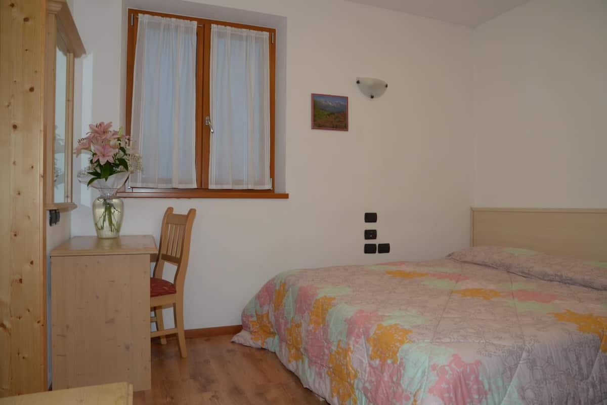Une chambre à coucher avec un lit, un bureau avec une chaise, une fenêtre avec des rideaux, un lampadaire fixé au mur et un petit tableau.