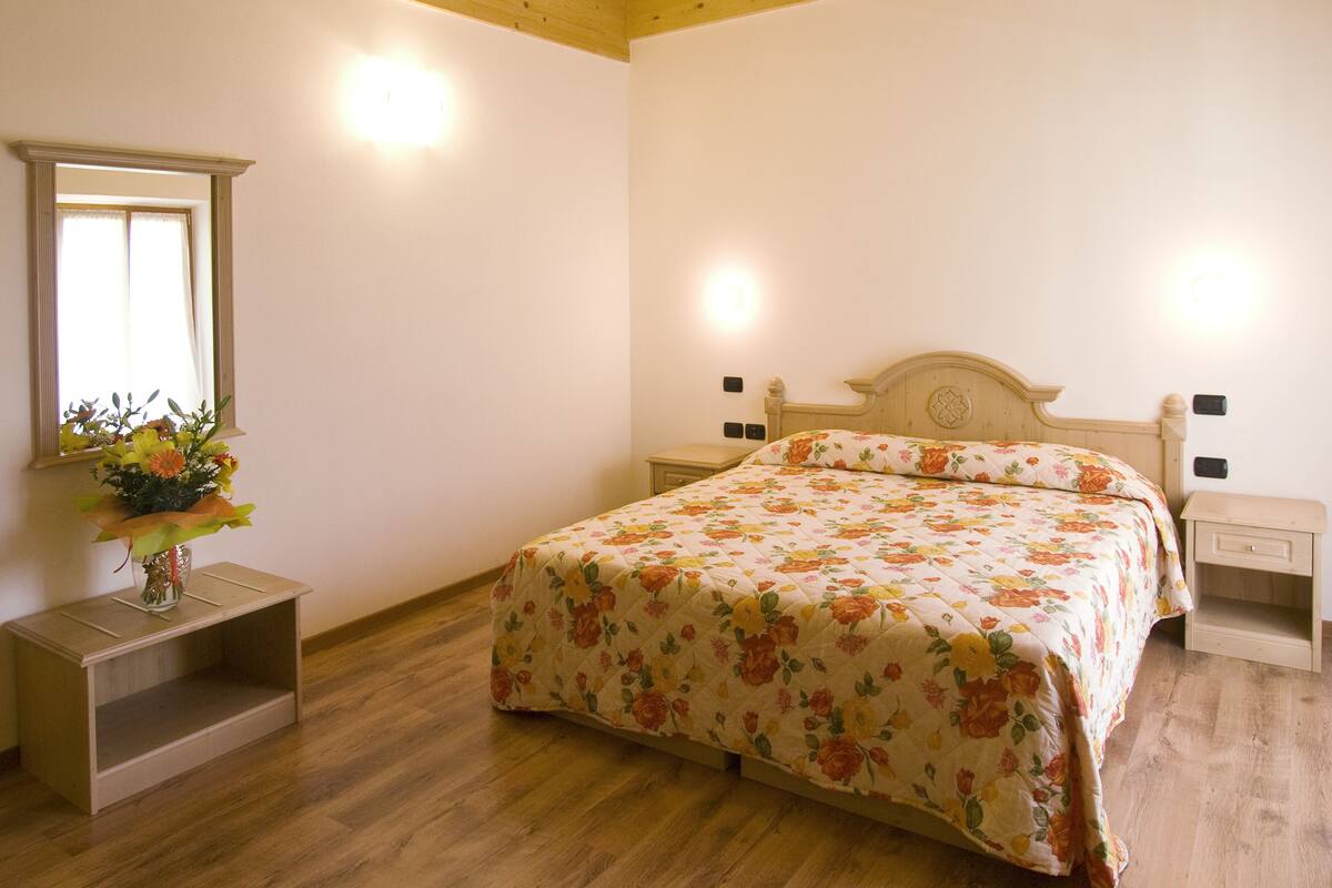Une chambre à coucher avec un lit en bois, une table de chevet, une commode, un miroir et un arrangement floral posé sur une étagère.