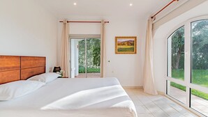 Une chambre avec un grand lit, une tête de lit en bois, une table de chevet avec une lampe et une vue sur un jardin, accessible par une porte coulissante.