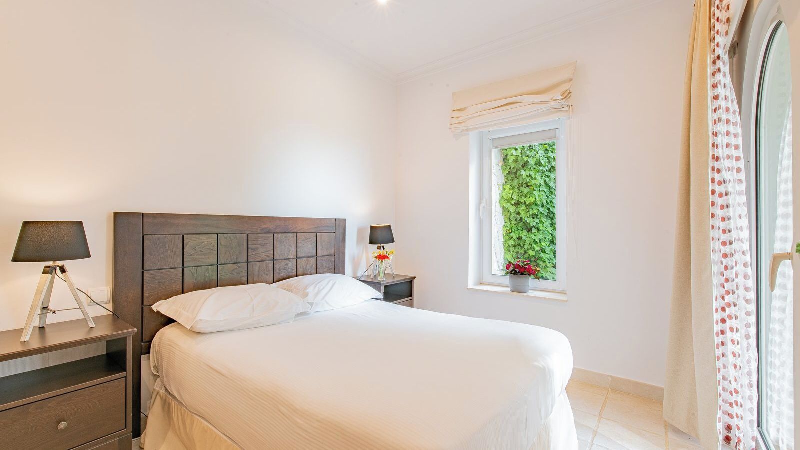 A neatly arranged bedroom with a bed, bedside tables, a lamp, a window with a view of greenery, and a curtain.