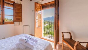 A bedroom with a bed, a wooden chair, and a balcony with a view of greenery and mountains.