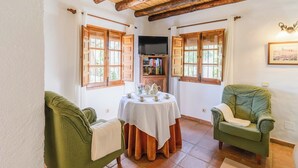 A cozy room with a round table, two green armchairs, wooden beams on the ceiling, and a bookshelf with books.