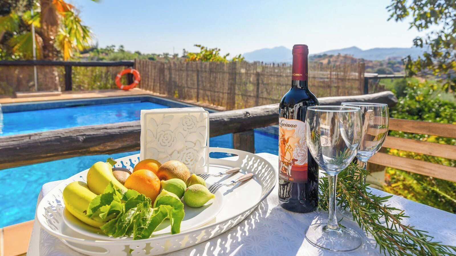 Ein Esstisch mit einer Fruchtschale, einer Weinflasche und zwei Gläsern, mit Blick auf einen Pool, eine Holzlattenveranda und Berge im Hintergrund.