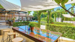 Un espace de restauration en plein air avec une table en verre et des tabourets, entouré de verdure et d’une piscine.