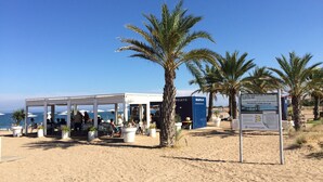 Ein Café am Strand mit Außenbestuhlung, Palmen und Blick auf das Meer.