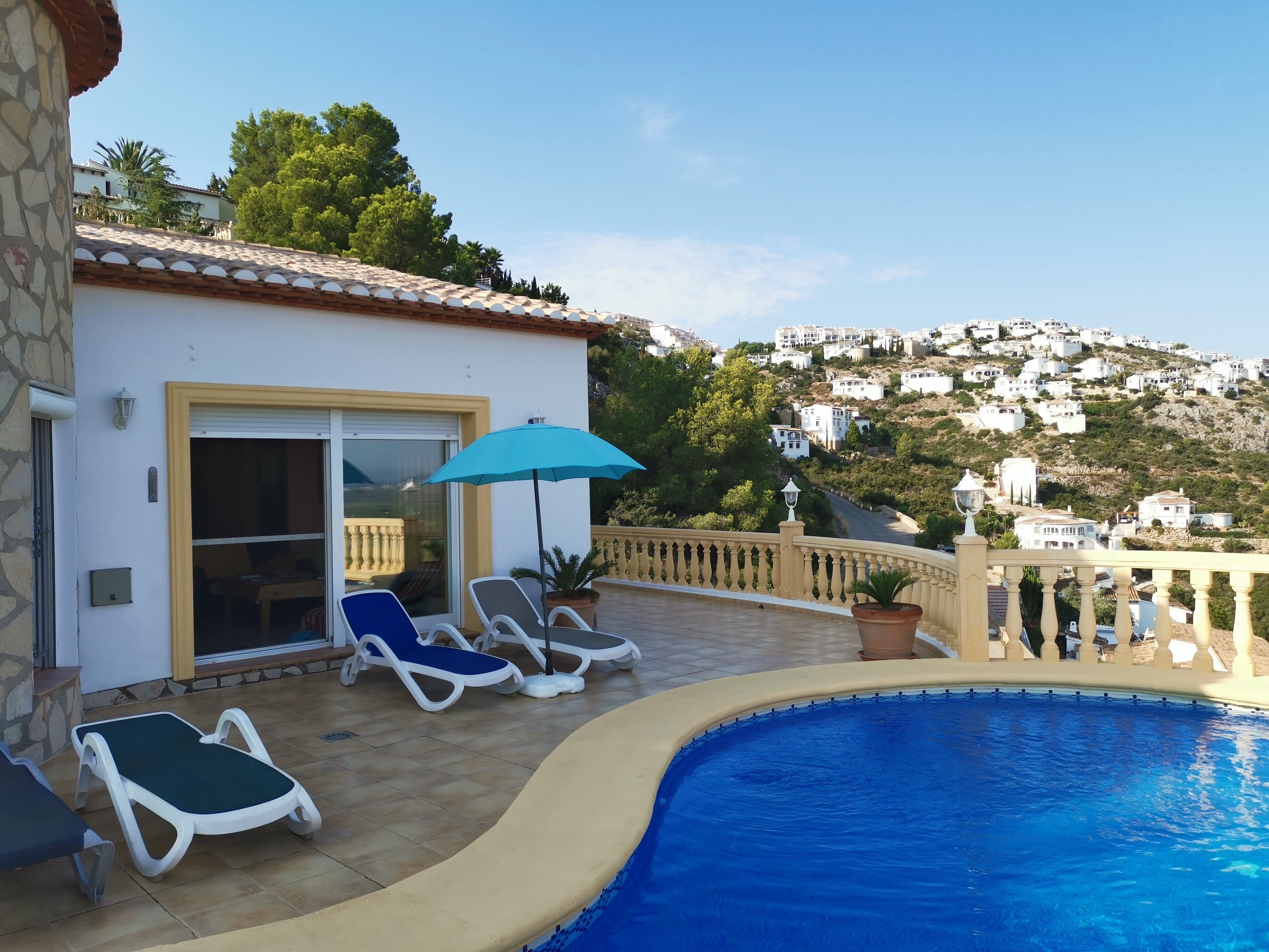 A pool area with lounge chairs and a blue umbrella, overlooking a hillside with white buildings.