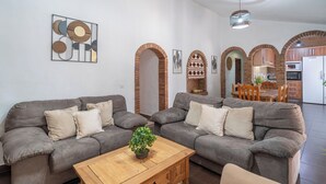 A living room with two grey sofas, a wooden coffee table, and a potted plant. There are brick arches and a kitchen in the background. - Herrero El Borge by Ruralidays (Málaga)
