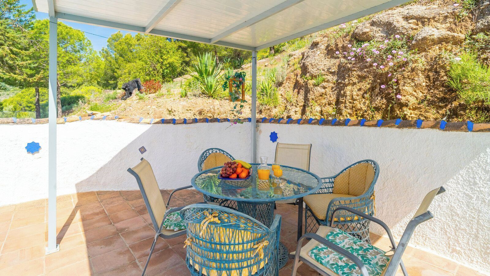 An outdoor patio with a round glass table, wicker chairs, and a view of a rocky hillside.