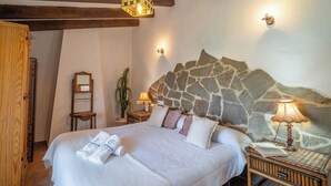 A bedroom with a stone headboard, a bed with white linens, two bedside lamps, and a wooden door. - Mulera Alozaina by Ruralidays (Málaga)