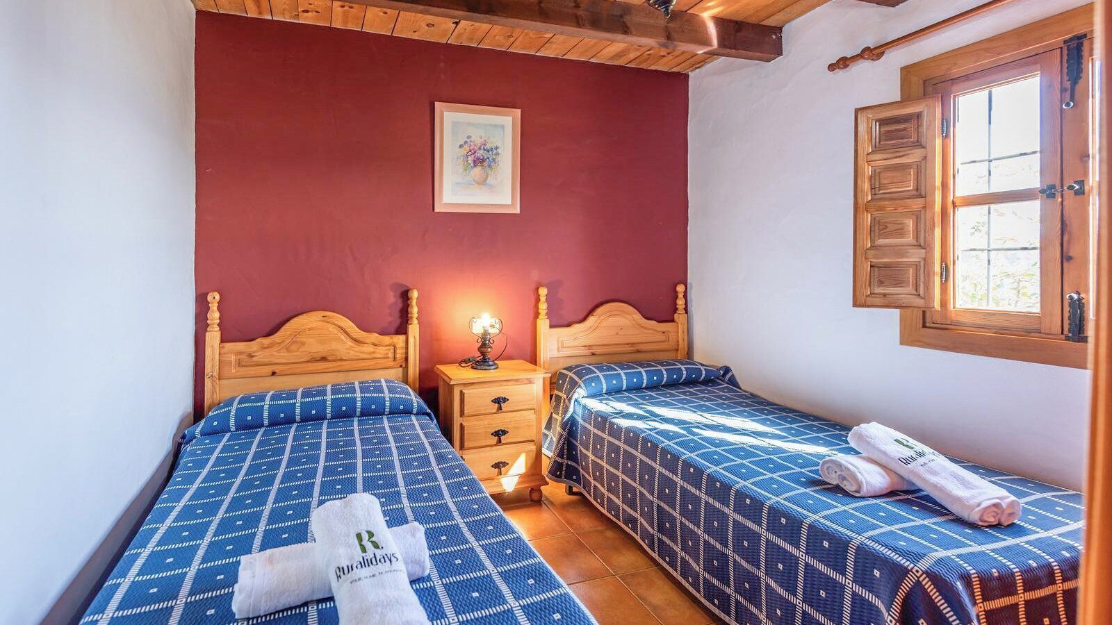 A bedroom with two beds, wooden headboards, a nightstand with a lamp, and a window with wooden shutters.