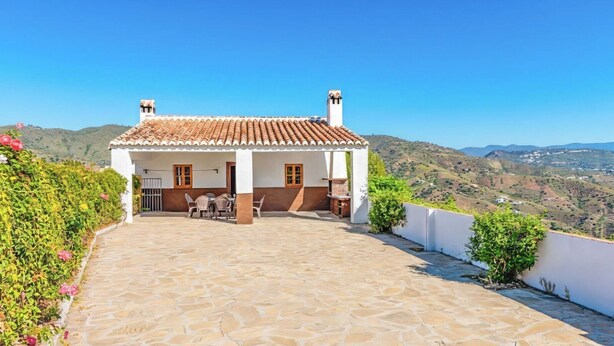 A white building with a tiled roof and a stone patio. There are two chimneys, a door, and a small table with chairs outside.