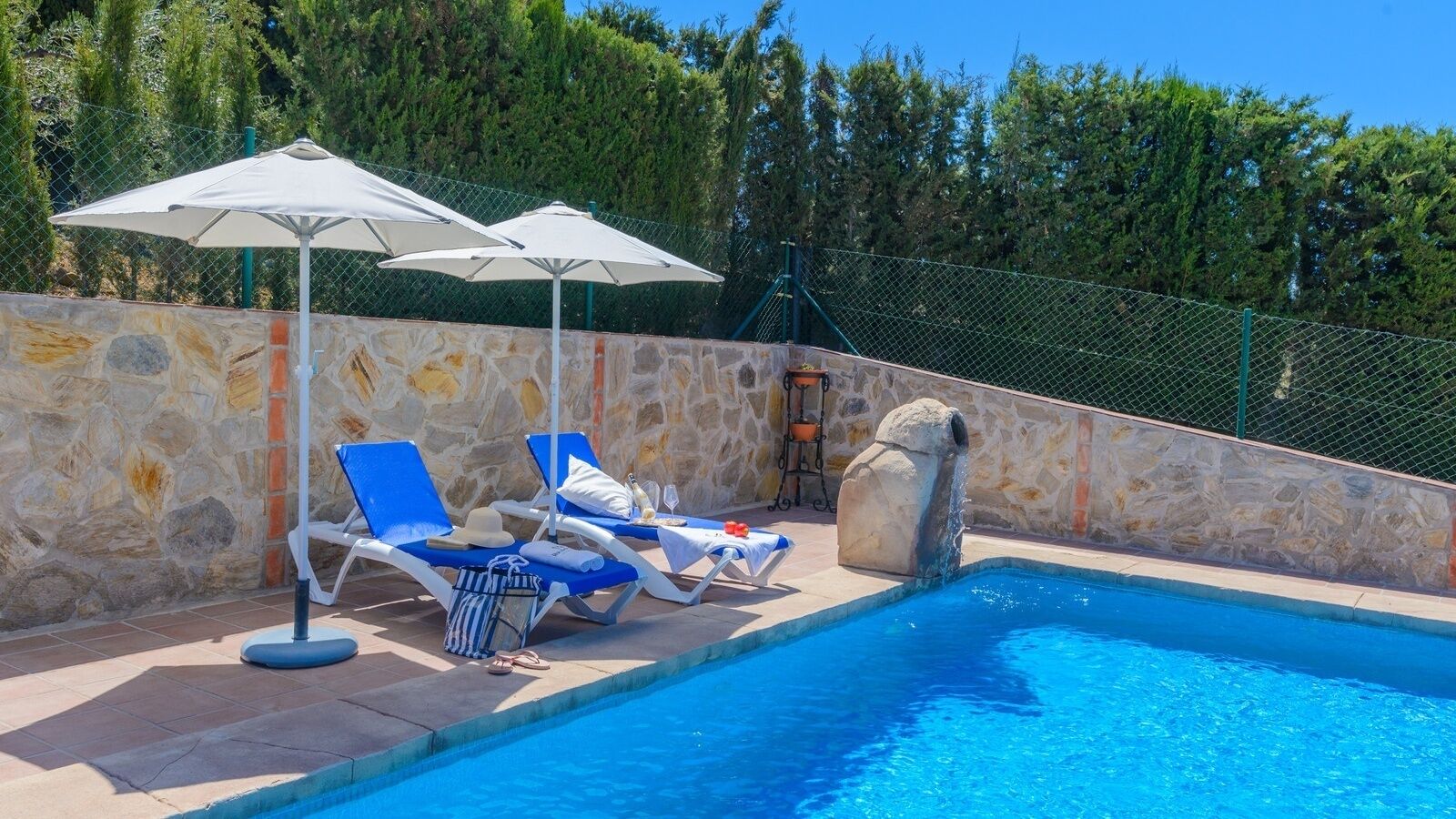 A swimming pool area with lounge chairs and umbrellas, surrounded by a stone wall and a fence.