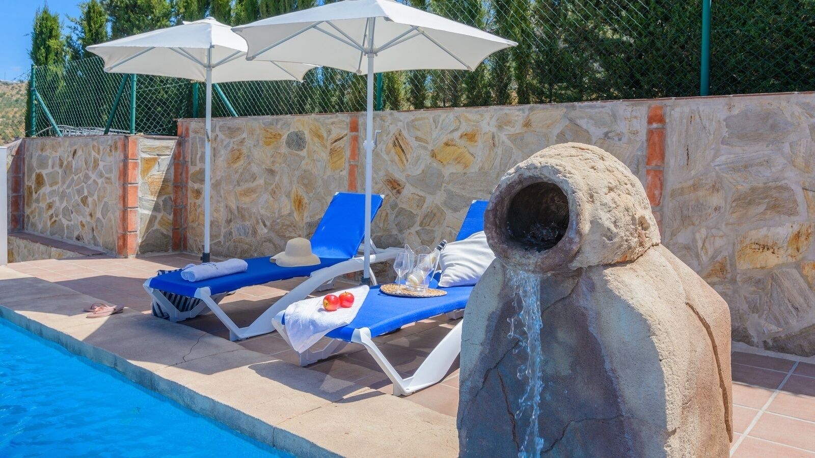 A pool area with stone walls, blue lounge chairs, and a stone structure with water flowing from it.