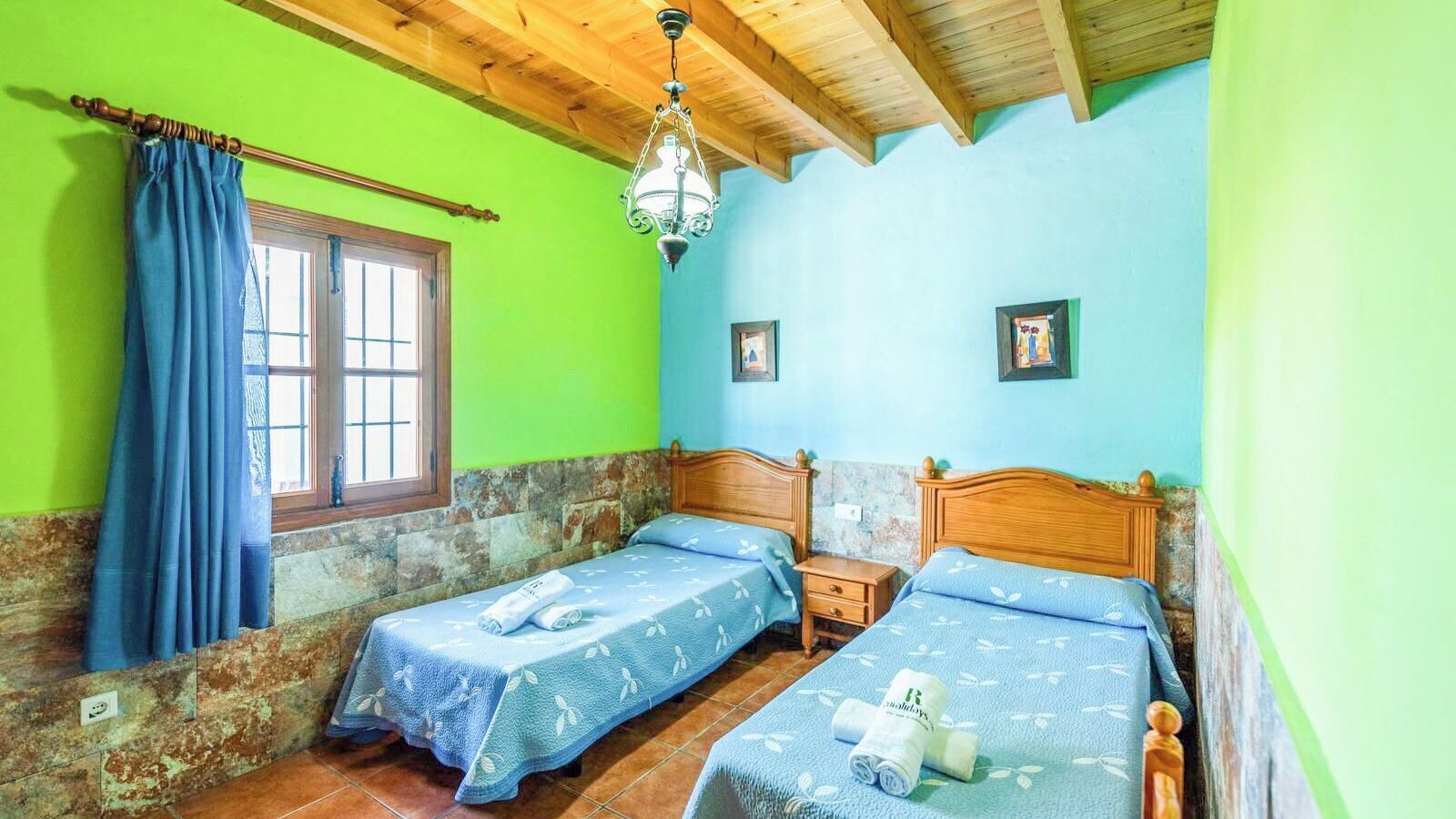 Une chambre avec deux lits, un plafond en bois, une fenêtre avec des rideaux bleus et un mur en pierre.