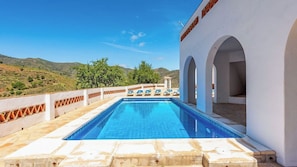 A swimming pool with a view of the countryside, a white building with arches, and a terrace with chairs.