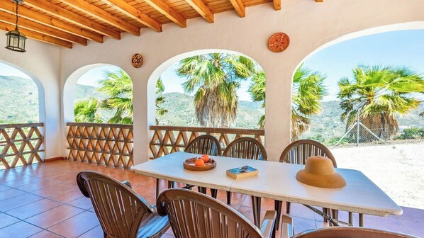 A covered outdoor dining area with a table, chairs, and a view of palm trees and hills.