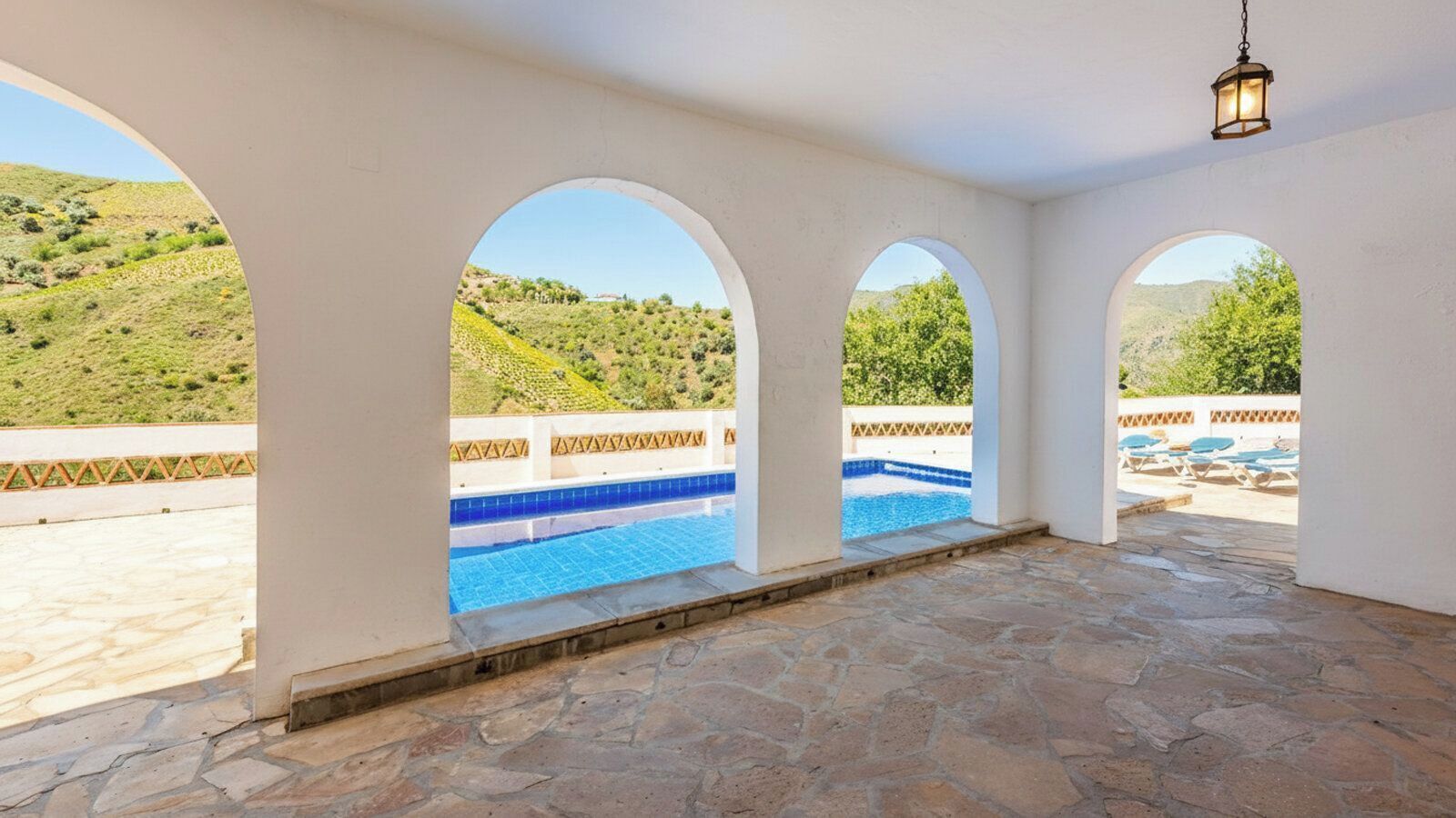 Ein überdachter Außenbereich mit Pool, Steinböden und bogenförmigen Fenstern mit Blick auf eine Hügellandschaft.