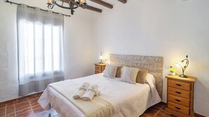 Cama bem arrumada com edredom branco e almofadas, duas mesas de cabeceira com abajures, cabeceira de madeira e uma janela com cortinas leves.