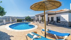 Piscina com espreguiçadeiras e um pérgola, rodeada por um edifício branco com telhado em azulejos.
