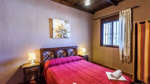 A bedroom with a wooden headboard, a red bedspread, two bedside lamps, a painting on the wall, a window with curtains, and a striped curtain on the right. - La Mina Benaoján by Ruralidays (Málaga)