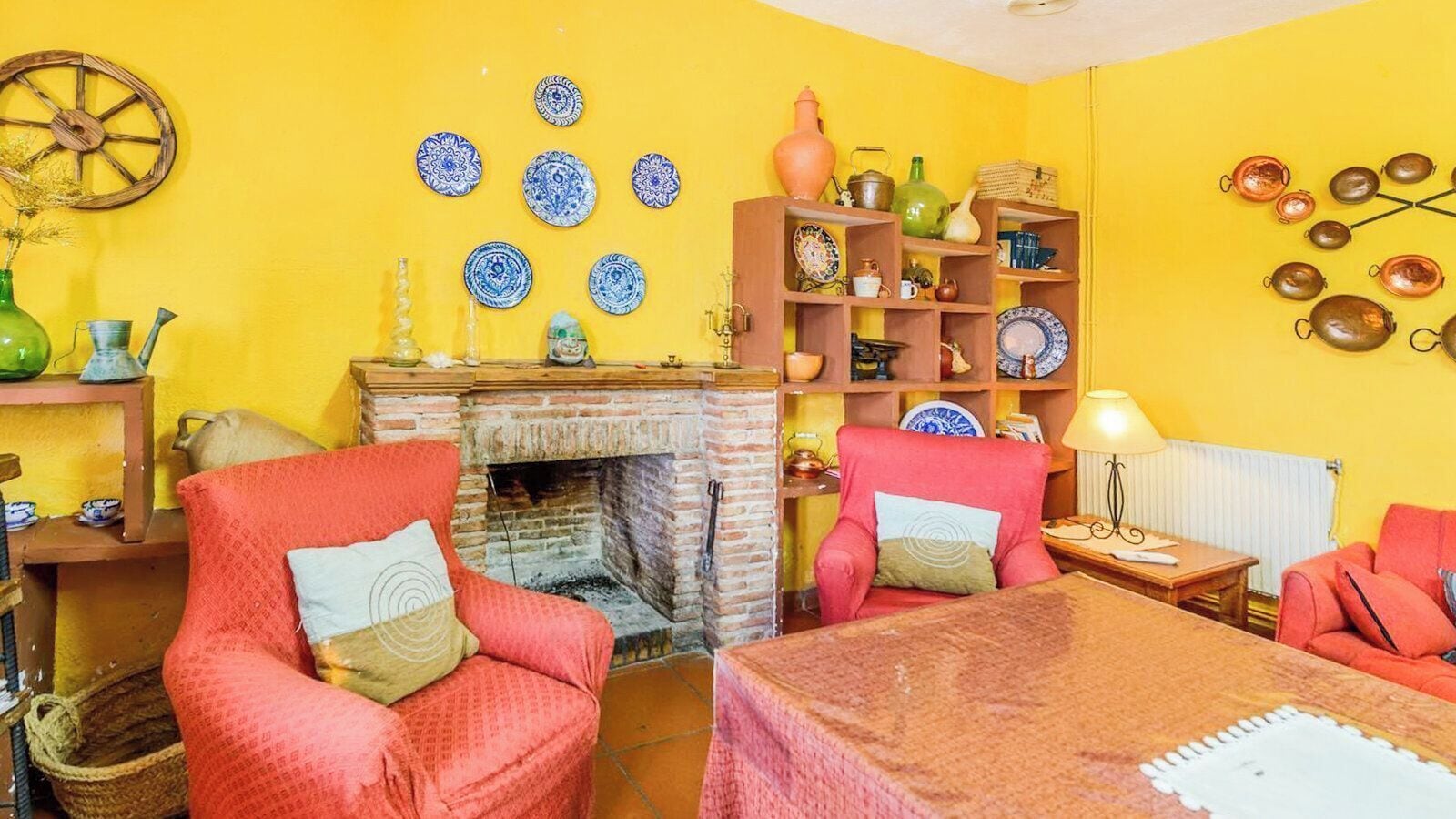 A room with a brick fireplace, two red armchairs, a wooden table, and a shelf with various decorative items.