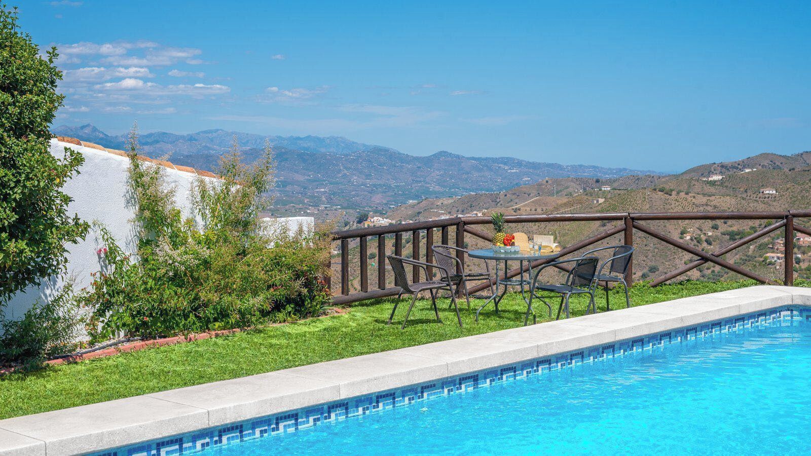 A swimming pool with a view of mountains, a table with chairs, and a wooden fence.