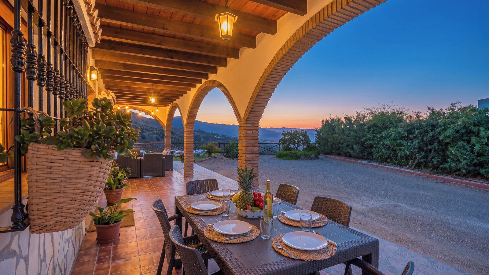An outdoor dining area with a table set for two, surrounded by wicker chairs, potted plants, and a view of the landscape.