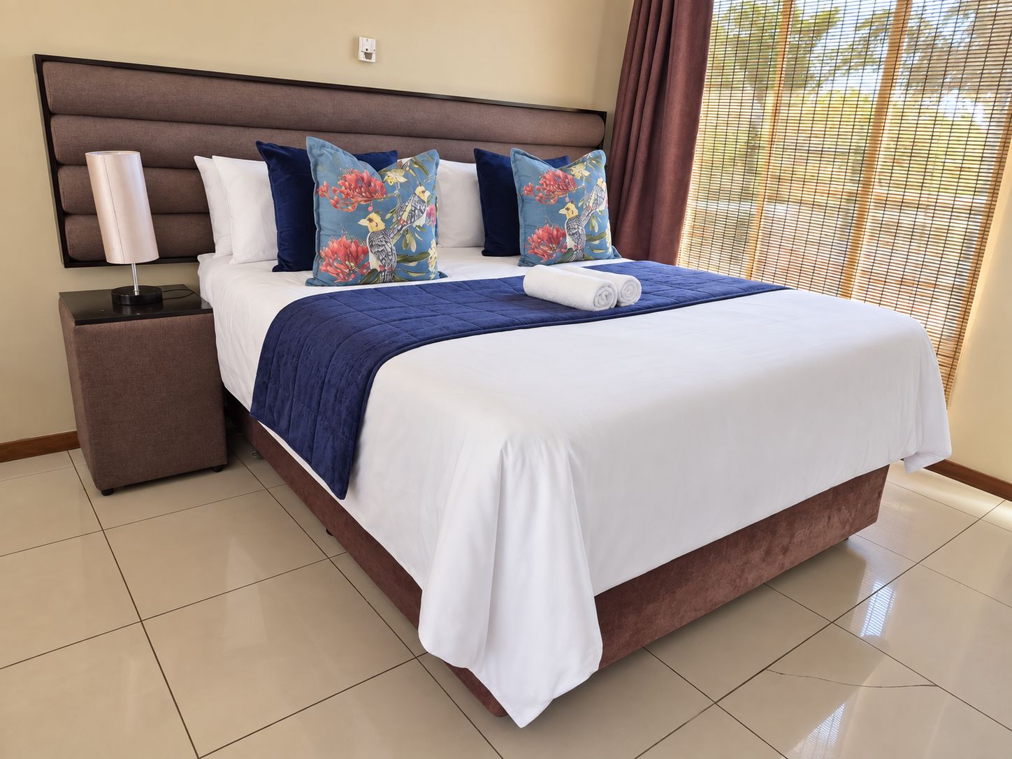 Una habitación de hotel con una cama grande, una mesita de noche, una lámpara y un ventanal con persianas.