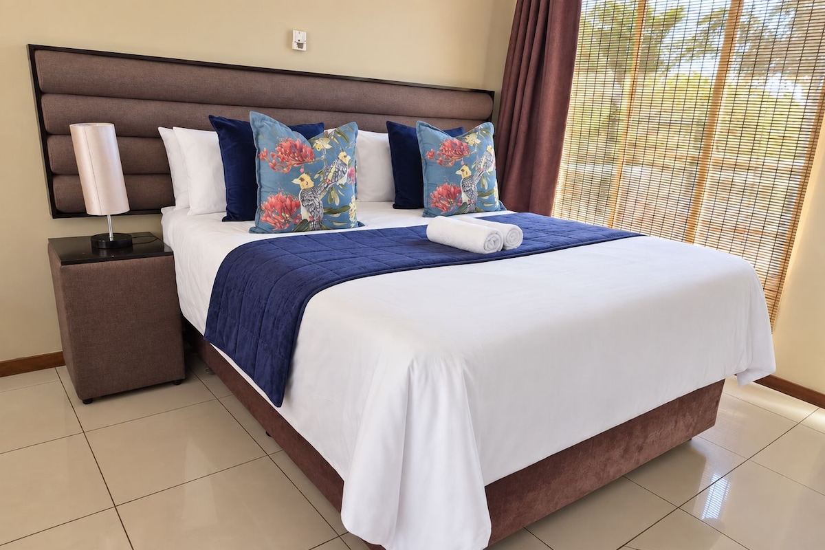 A hotel room with a large bed, a nightstand, a lamp, and a window with blinds.