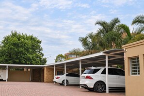 A covered parking area with two white cars parked under a shelter, surrounded by trees and a building with a brick facade. - Croydon hotel (Kempton Park)