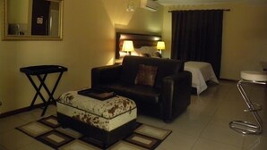 A hotel room with a sofa, a small table, a chair, a bed with a white cover, a mirror, and an air conditioning unit. - Croydon hotel (Kempton Park)