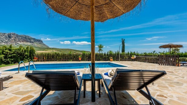 Un área de piscina con sillas de descanso y una mesa, rodeada por una valla de madera y plantas en macetas.