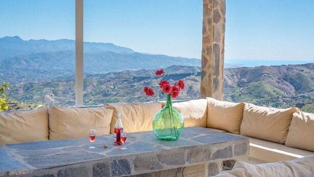 Una mesa de piedra con un jarrón de flores rojas, dos vasos de líquido rojo y una botella, con una vista panorámica de montañas al fondo.