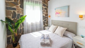 Una cama bien hecha con dos almohadas y dos toallas dobladas, una planta en maceta, una pared de piedra y un cuadro colgado.
