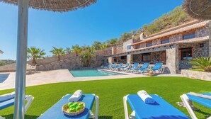 Un área junto a la piscina con sillones de descanso, una piscina y un edificio de piedra con tejado de baldosas.