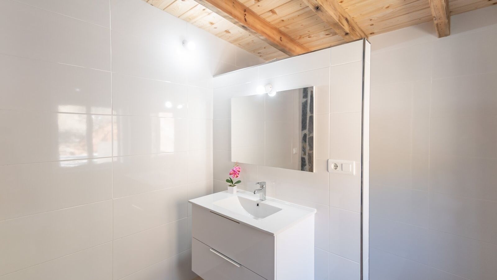 A modern bathroom with white tiles, a white sink, a mirror, and a wooden ceiling.