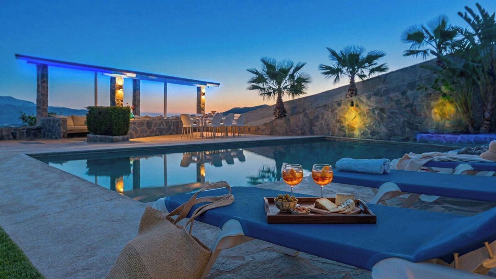 A poolside area with lounge chairs, a dining setup with glasses and snacks, and a stone wall backdrop.