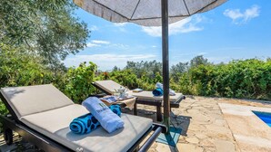Zona de estar al aire libre iluminada por el sol, con sillones, una mesa con una toalla y una taza, y una piscina al fondo.