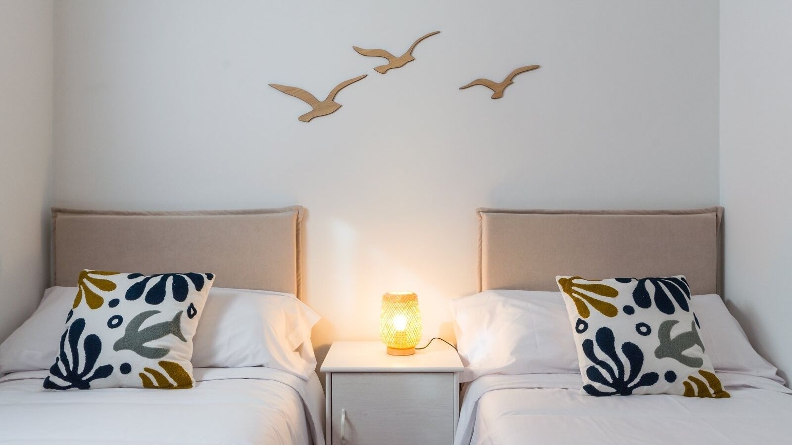 Et hotelværelse med to senge, hver med et mønstret pude, et natbord med en lampe og billeder af fugle på væggen.
