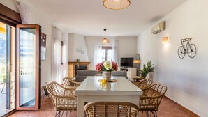 A dining area with a glass table, wicker chairs, a vase with flowers, a fireplace, a television, and a bicycle mounted on the wall. - Cortijo Angela Viñuela by Ruralidays (Málaga)