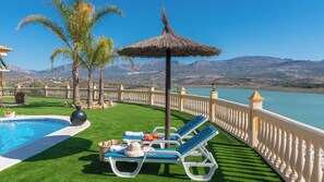 A poolside area with a thatched umbrella, lounge chairs, and a view of a lake and mountains. - Cortijo Angela Viñuela by Ruralidays (Málaga)