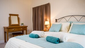 Una cama bien hecha con colcha azul y blanca, una mesita de noche de madera con lámpara, un espejo y una pequeña mesa con una planta.